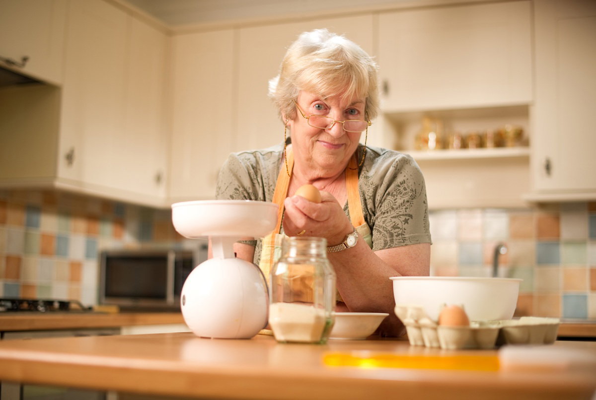 kobieta w wieku 60 plus w okularach i fartuszku przygotowuje jedzenie w kuchni i lekko się uśmiecha