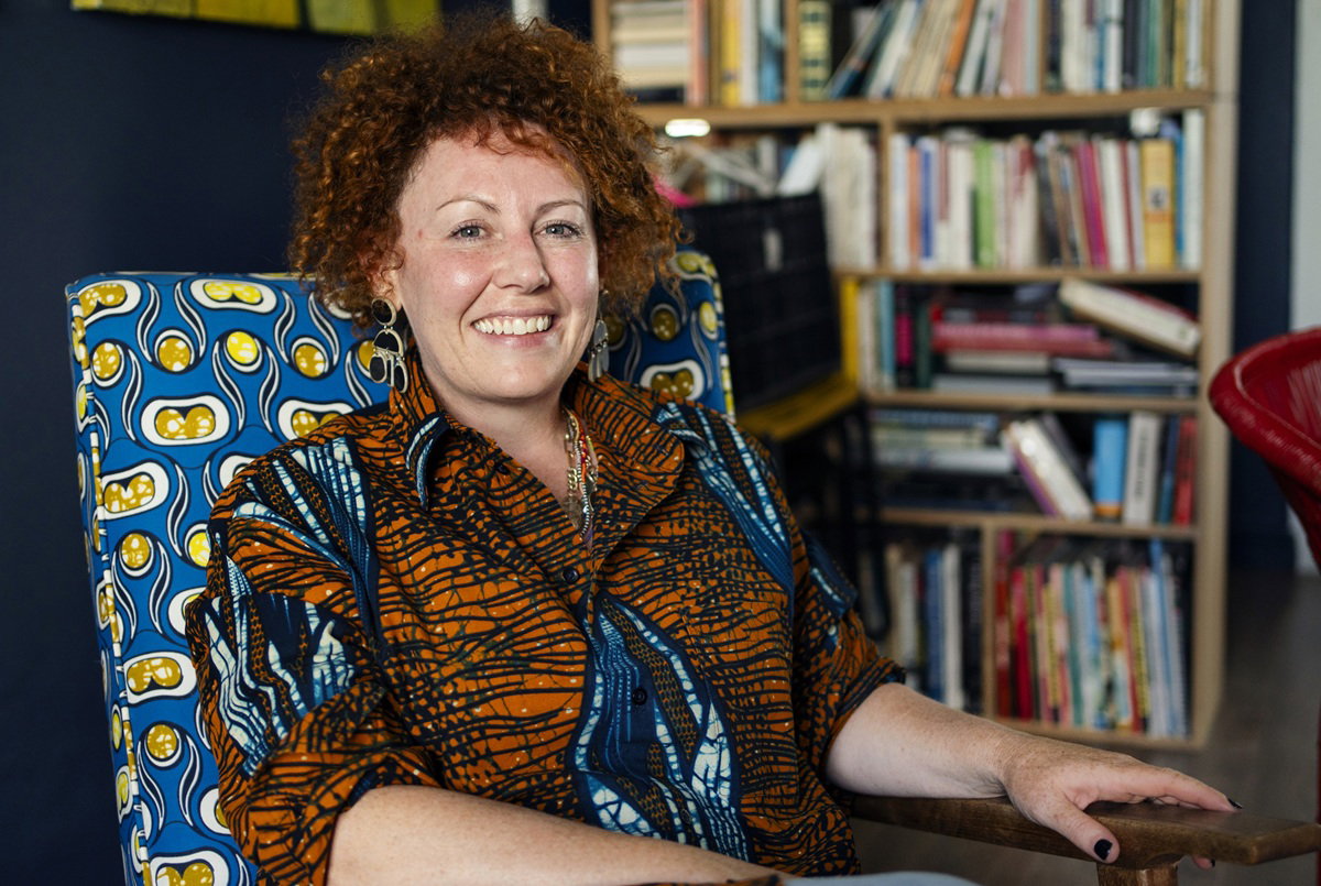 kobieta w wieku 40 plus siedzi w fotelu w stylu 60 plus na tle półki z książkami i się uśmiecha