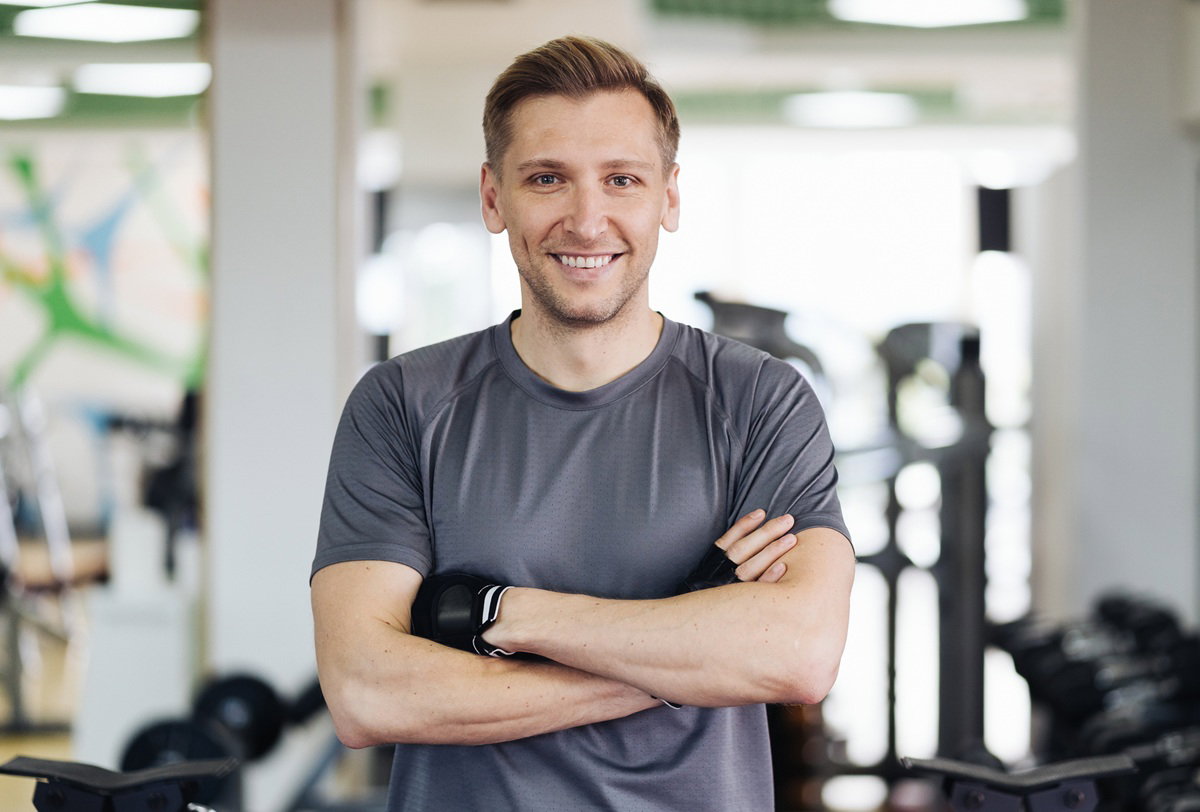 mężczyzna w wieku 35 plus w szarej koszulce na sali treningowej uśmiecha się zadowolony