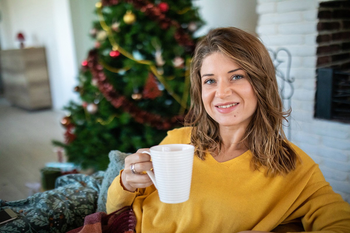 kobieta w wieku 25 plus siedzi na tle choinki w żółtym swetrze z kubkiem herbaty i się uśmiecha
