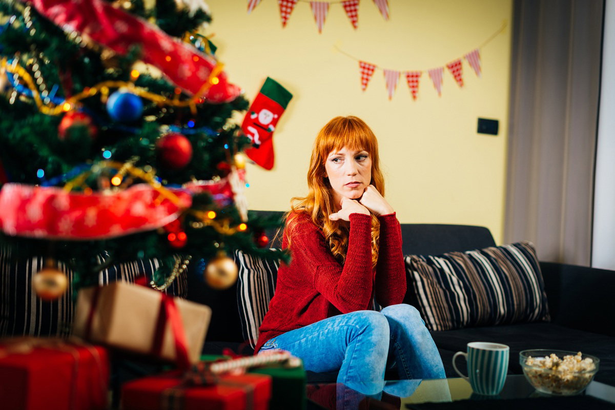 kobieta w wieku 30 plus siedzi an kanapie i patrzy smutno w stronę choinki