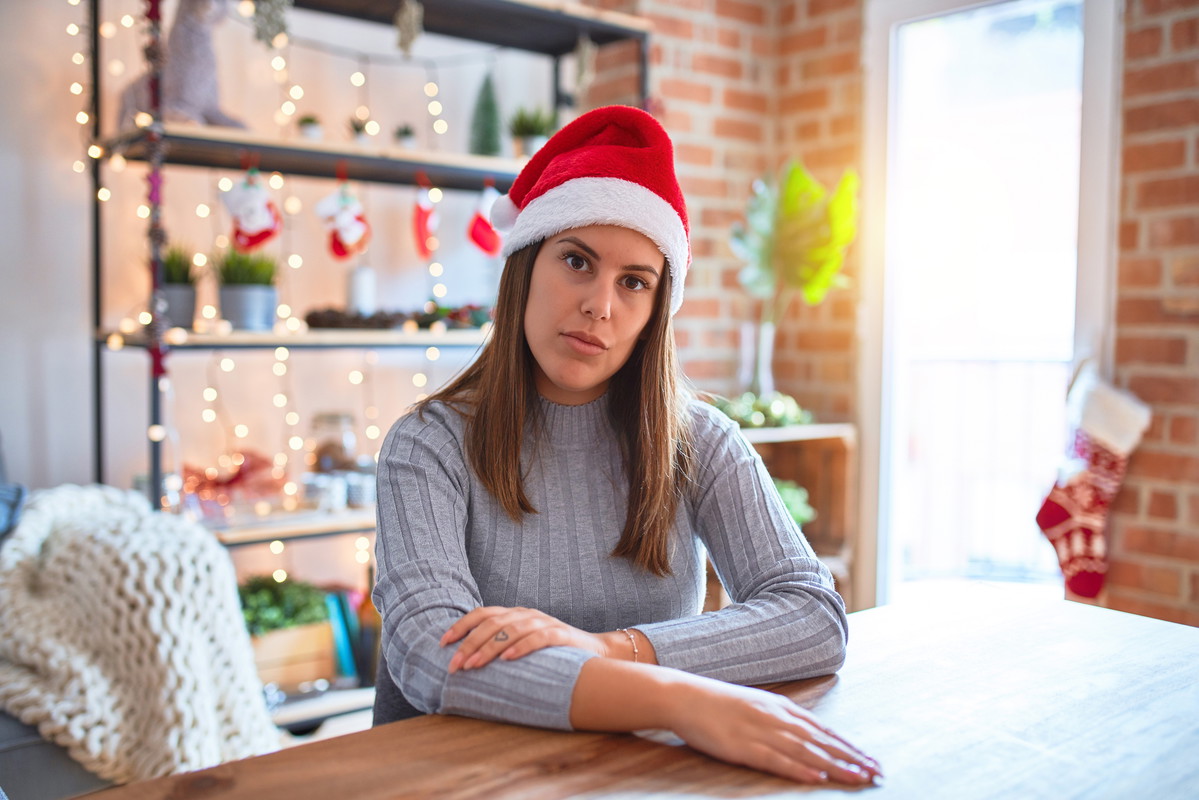 Young beautiful woman wearing christmas hat sitting at the table at home with serious expression on face. Simple and natural looking at the camera.