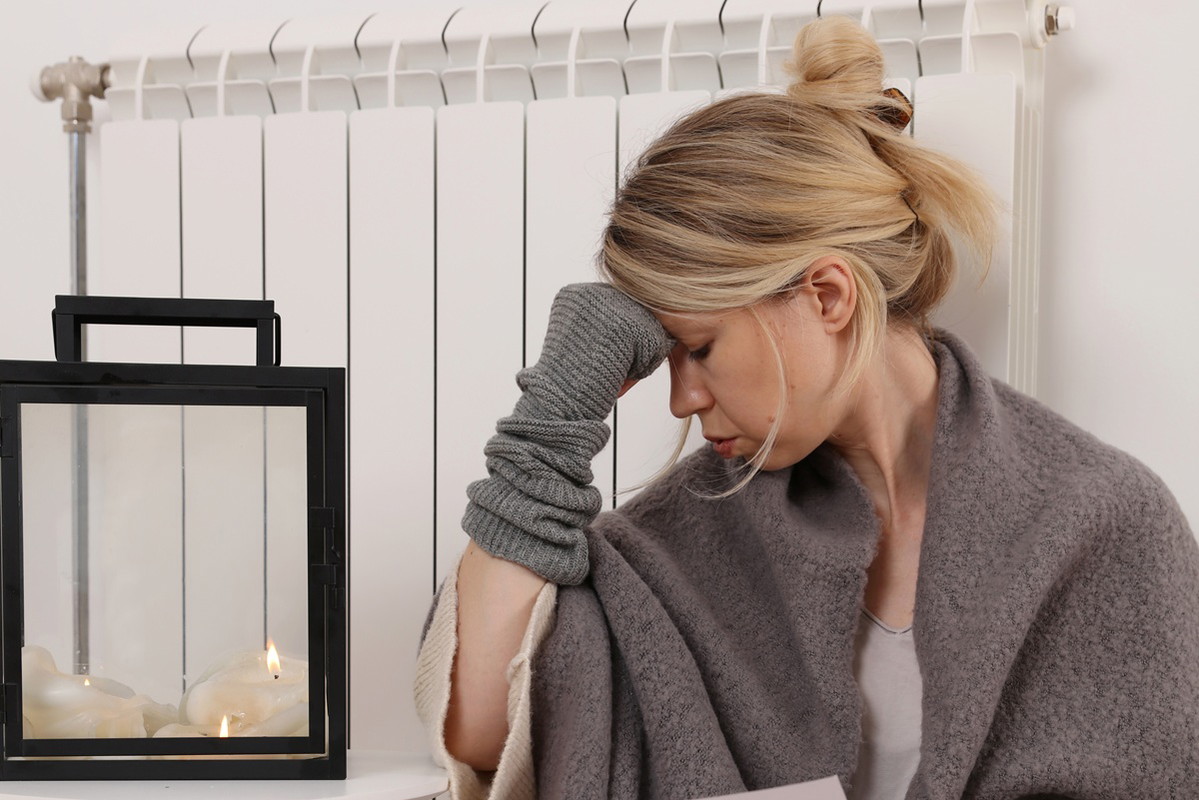 kobieta w wieku 35 plus siedzi w kocu i rękawiczkach przy zimnym kaloryferze i patrzy na rachunki