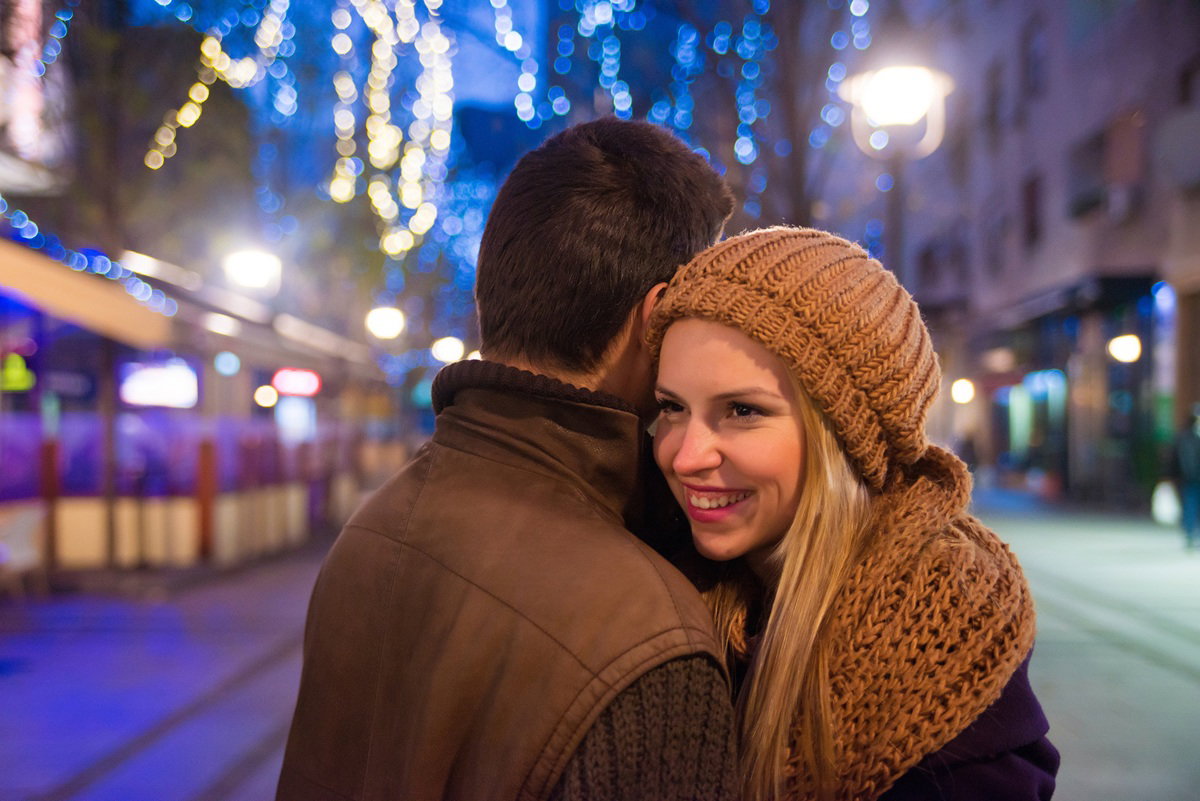 kobieta w wieku 30 plus w karmelowej czapce i szalu uśmiecha się w ramionach mężczyzny na ulicy