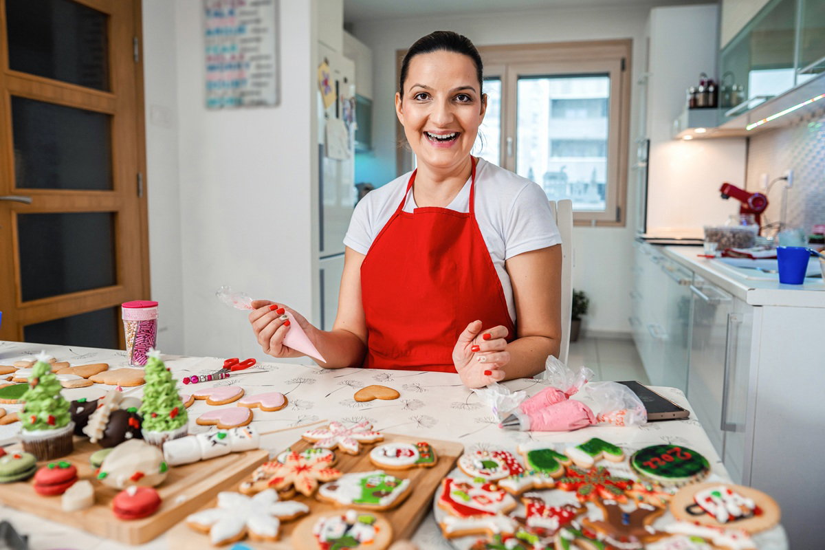kobieta w wieku 30 plus w czerwonym fartuszku w kuchni ozdabia świąteczne pierniczki