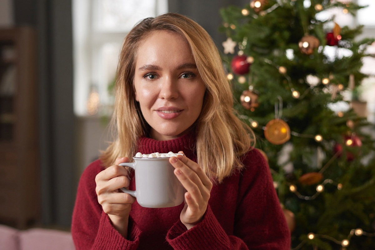 kobieta w wieku około 30 plus z kubkiem gorącej czekolady z piankami siedzi na tle choinki