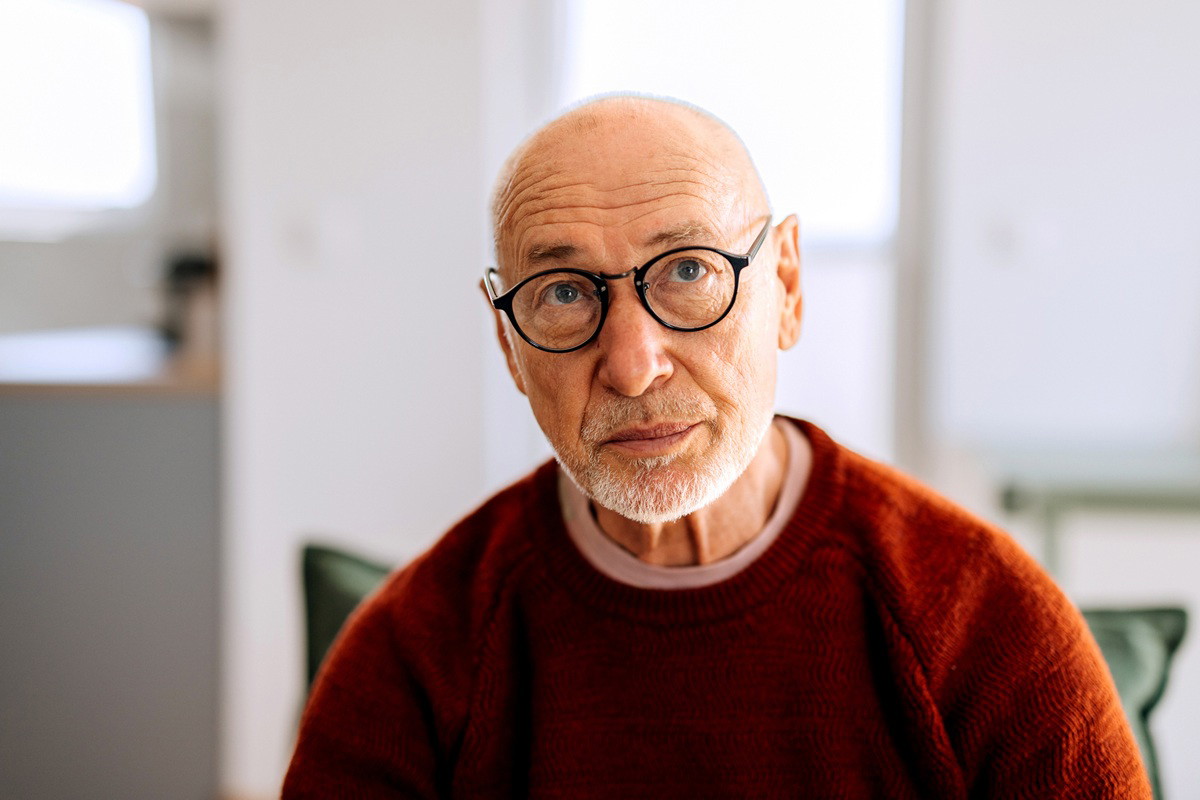 mężczyzna w wieku około 70 lat z siwymi zarostem w okularach siedzi na kanapie zmartwiony