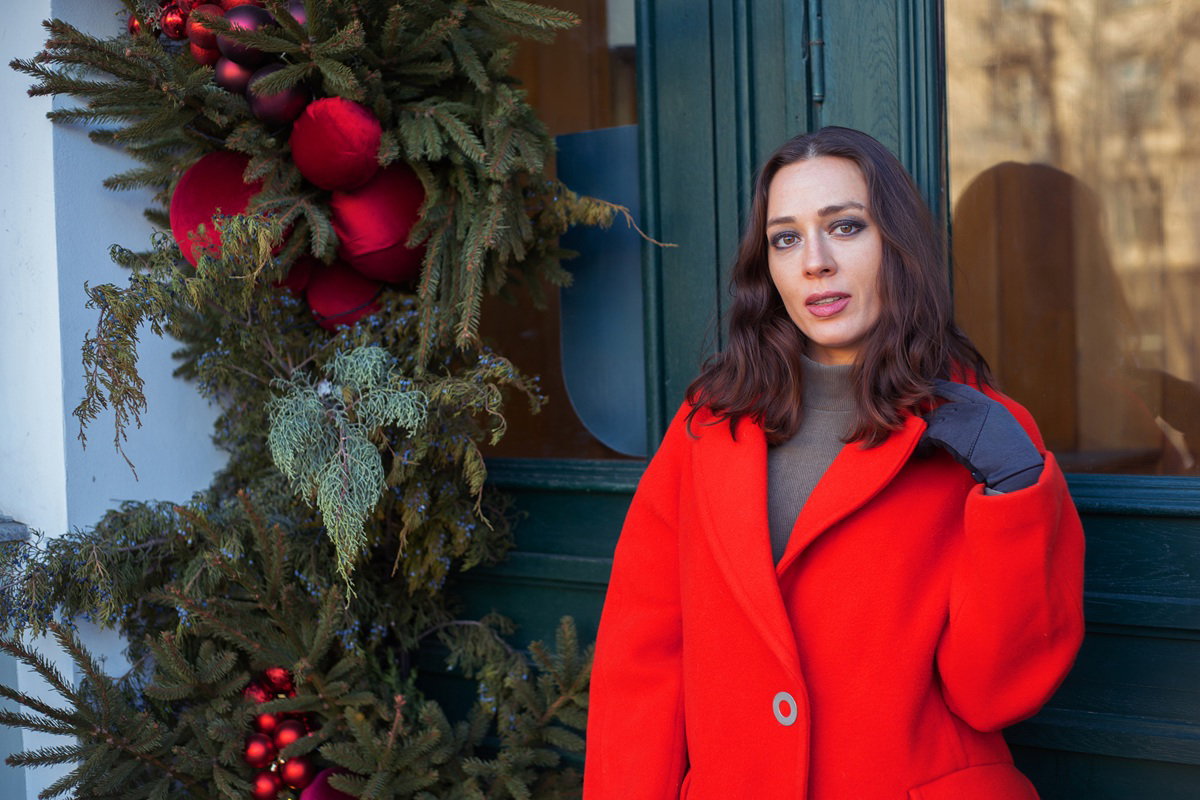 kobieta w wieku 30 plus w czerwonym płaszczu patrzy z triumfem stojąc na tle drzwi ozdobionych świątecznie
