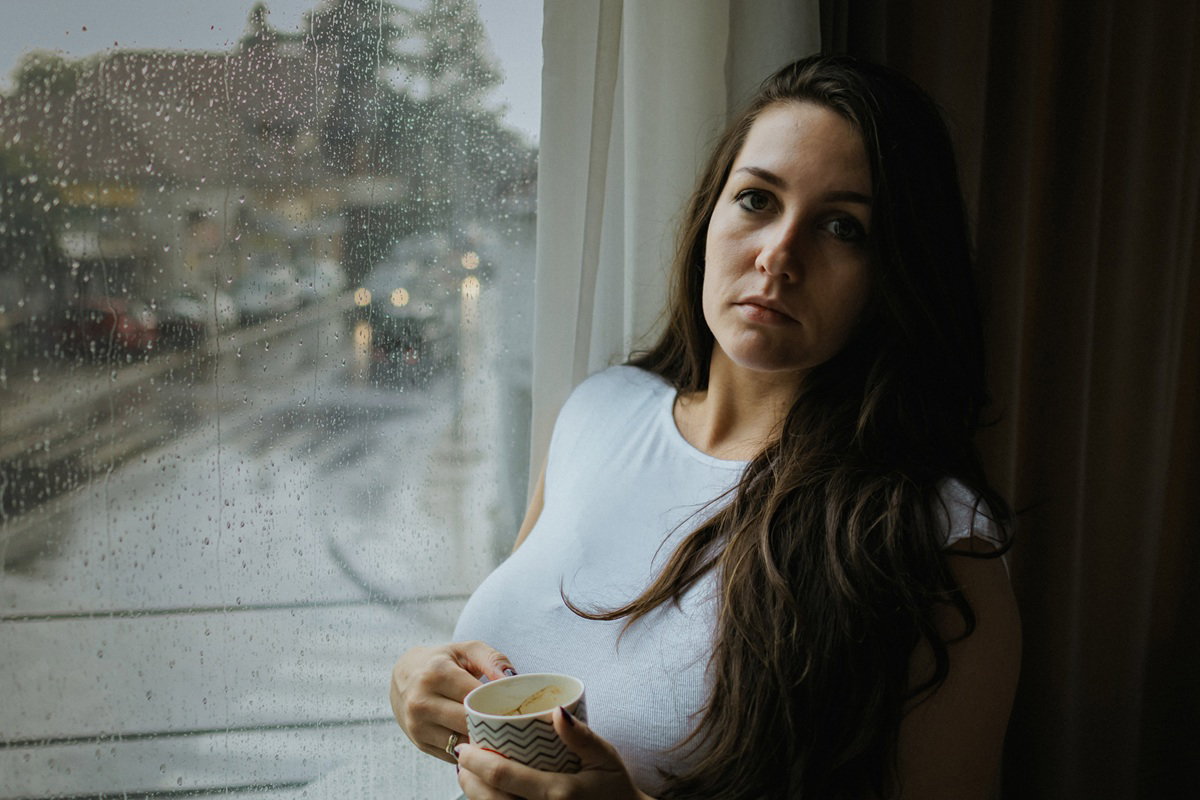 kobieta w wieku 30 plus stoi przy oknie z kubkiem kawy zrozpaczona a na ulicy pada deszcz