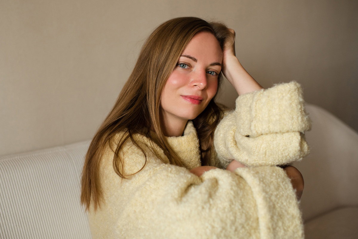 kobieta w wieku 30 plus w masełkowym swetrze siedzi na kanapie i lekko się uśmiecha