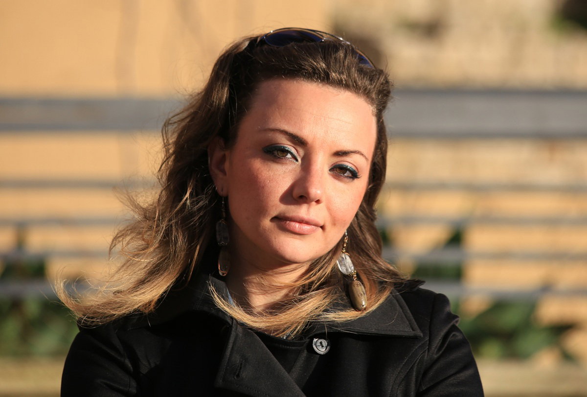 kobieta w wieku 35 plus stoi na zewnątrz w płaszczu w słońcu patrzy poważnym spojrzeniem