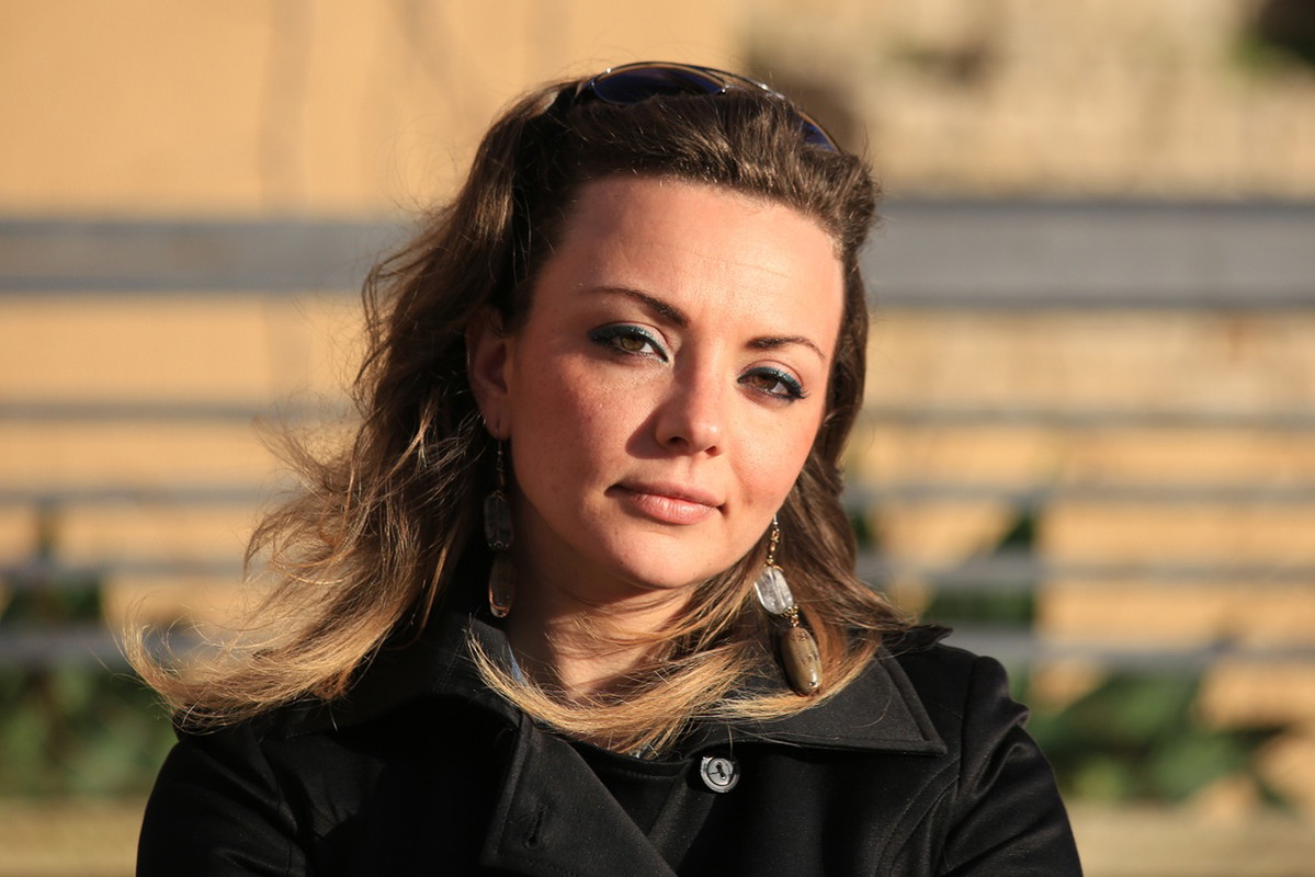 kobieta w wieku 35 plus stoi na zewnątrz w płaszczu w słońcu patrzy poważnym spojrzeniem