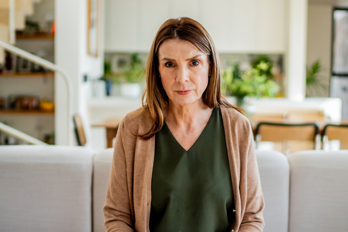 kobieta w wieku 60 plus w beżowym sweterku siedzi na kanapie zasmucona i poważna