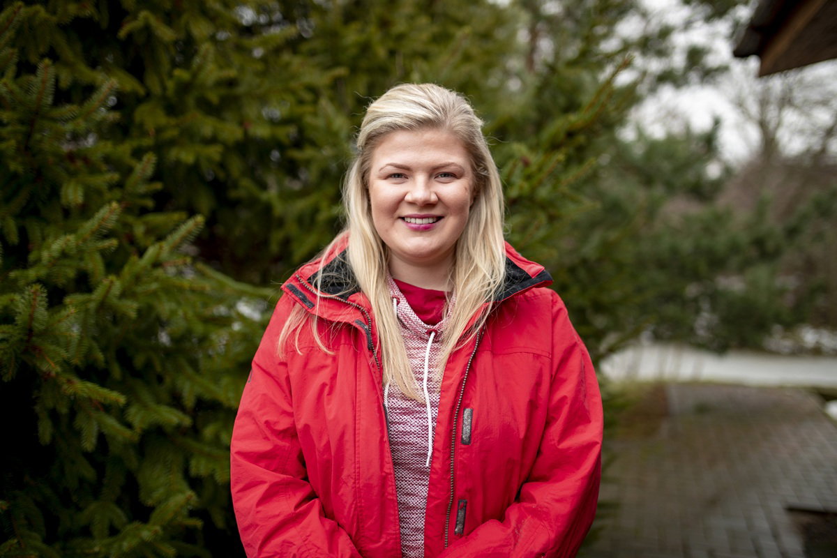 kobieta w wieku 30 plus w czerwonej kurtce blondynka stoi na tle zieleni i się uśmiecha