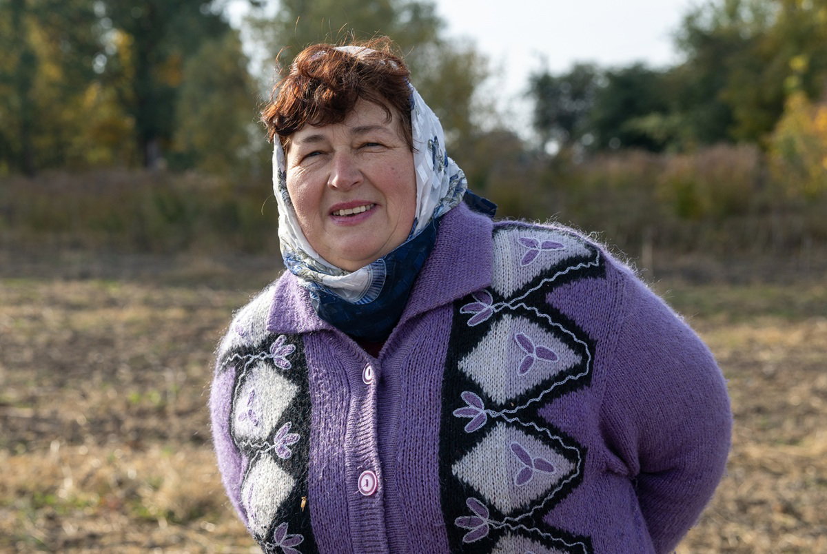 kobieta w wieku około 60 lat w chuście na głowie i we wzorzystym swetrze na guziki stoi na polu i lekko się uśmiecha