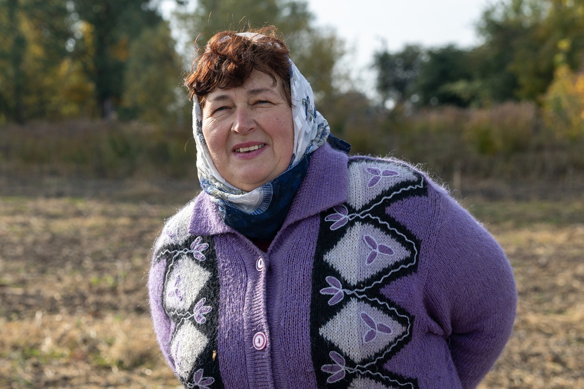 kobieta w wieku około 60 lat w chuście na głowie i we wzorzystym swetrze na guziki stoi na polu i lekko się uśmiecha