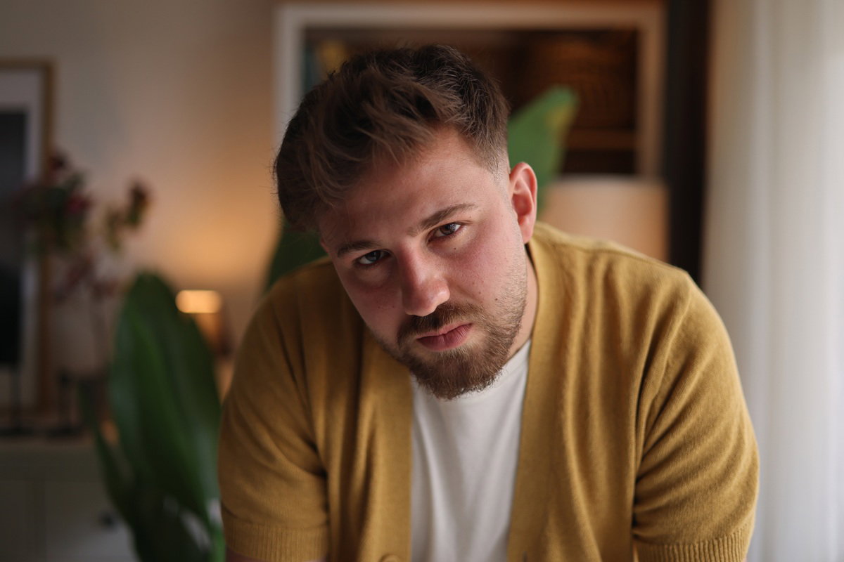 mężczyzna w wieku 20 plus z wąsami i brodą w żółtym swetrze patrzy smutny i zamyślony przed siebie