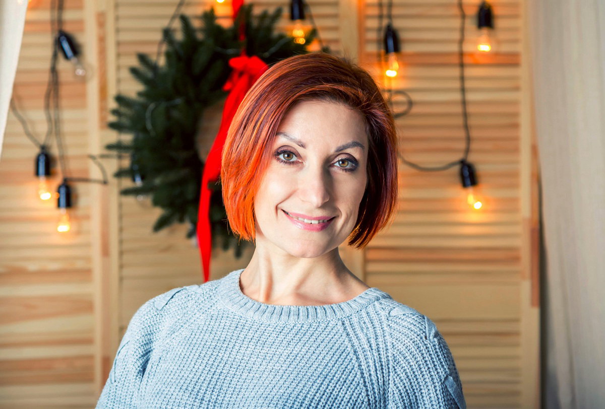 kobieta w wieku 40 plus w szarym sweterku na tle drewnianej szafy i wieńca świątecznego uśmiecha się zażenowana