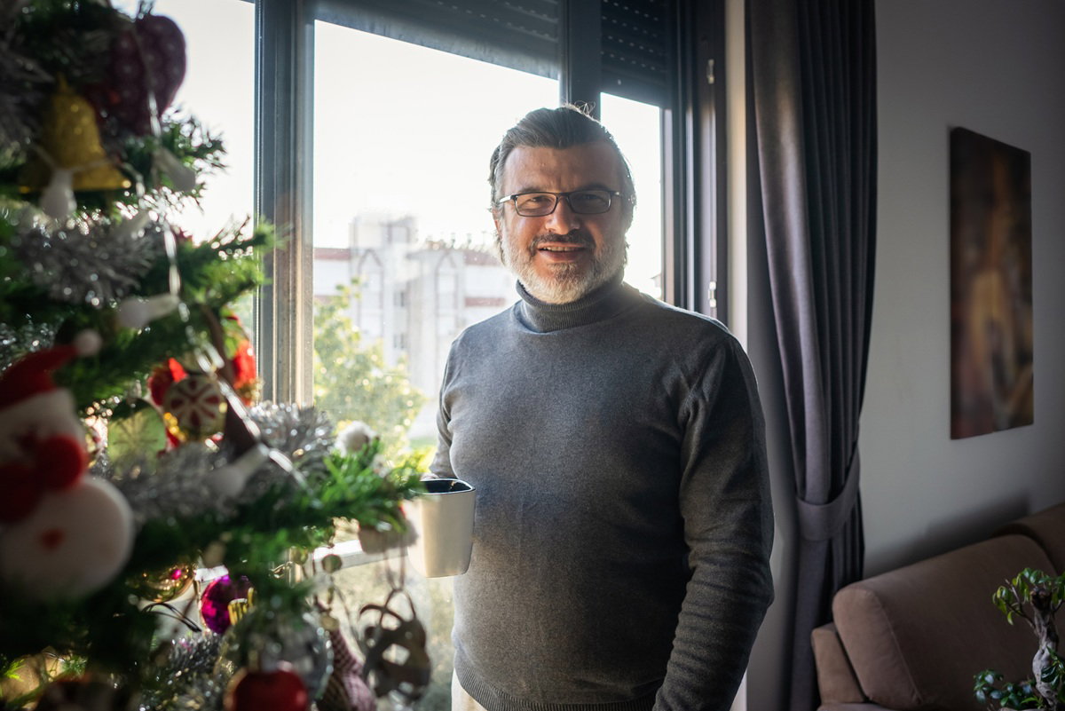 mężczyzna w wieku około 50 plus w szarym golfie i okularach stoi przy oknie i choince z kubkiem herbaty