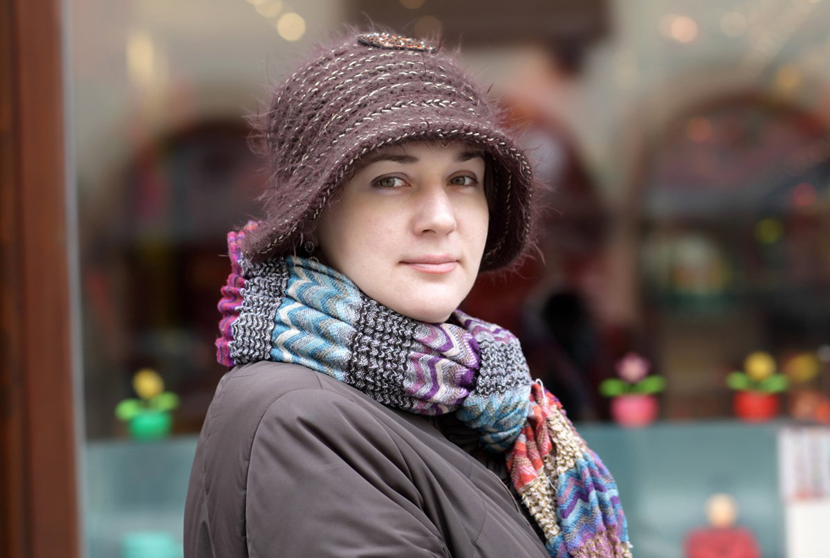 kobieta w wieku 30 plus w kapeluszu i kolorowym szalu na szyi stoi przed witryną sklepową zamyślona