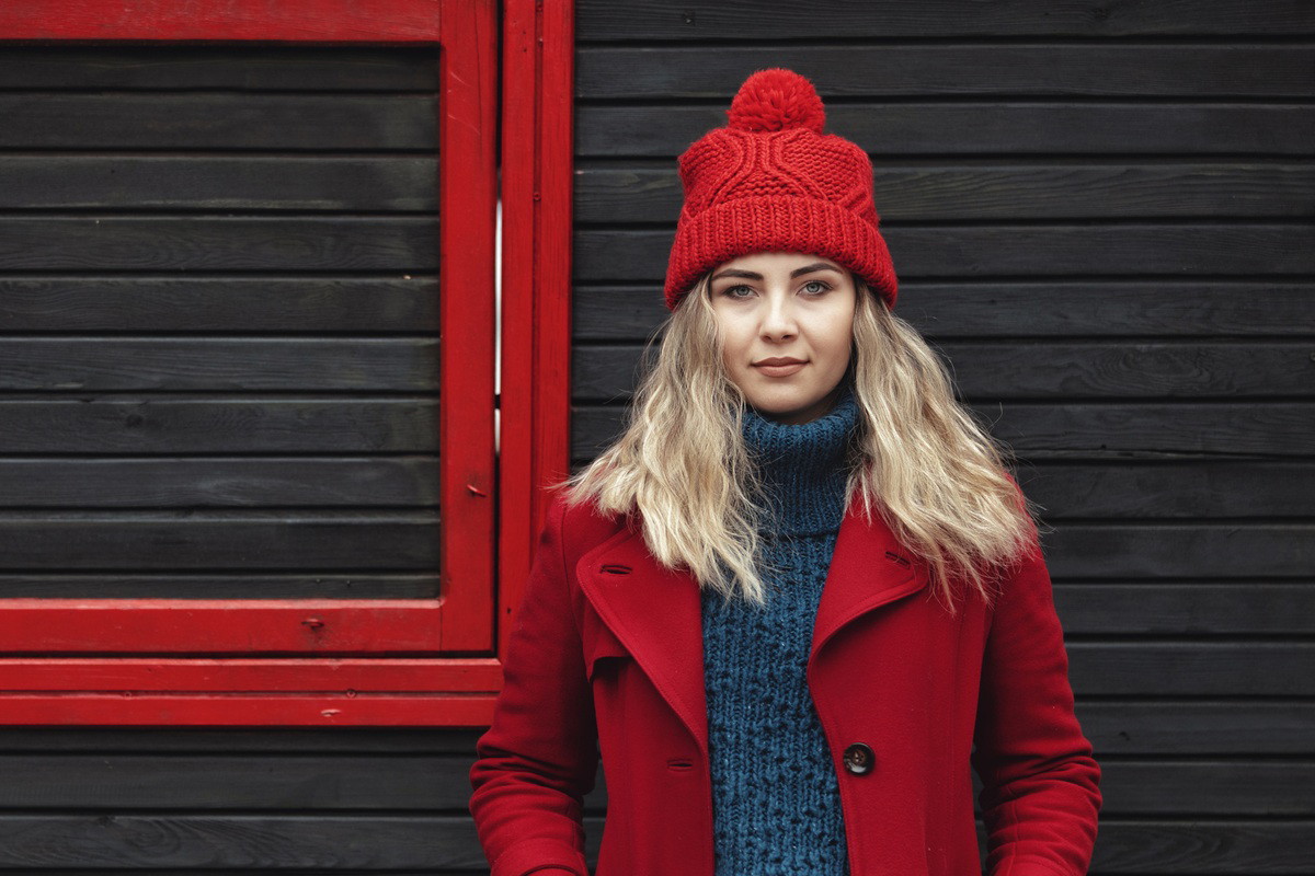 kobieta w wieku 25 plus w czerwonym płaszczu i czapce stoi na tle drewnianej ściany z poważnym wyrazem twarzy