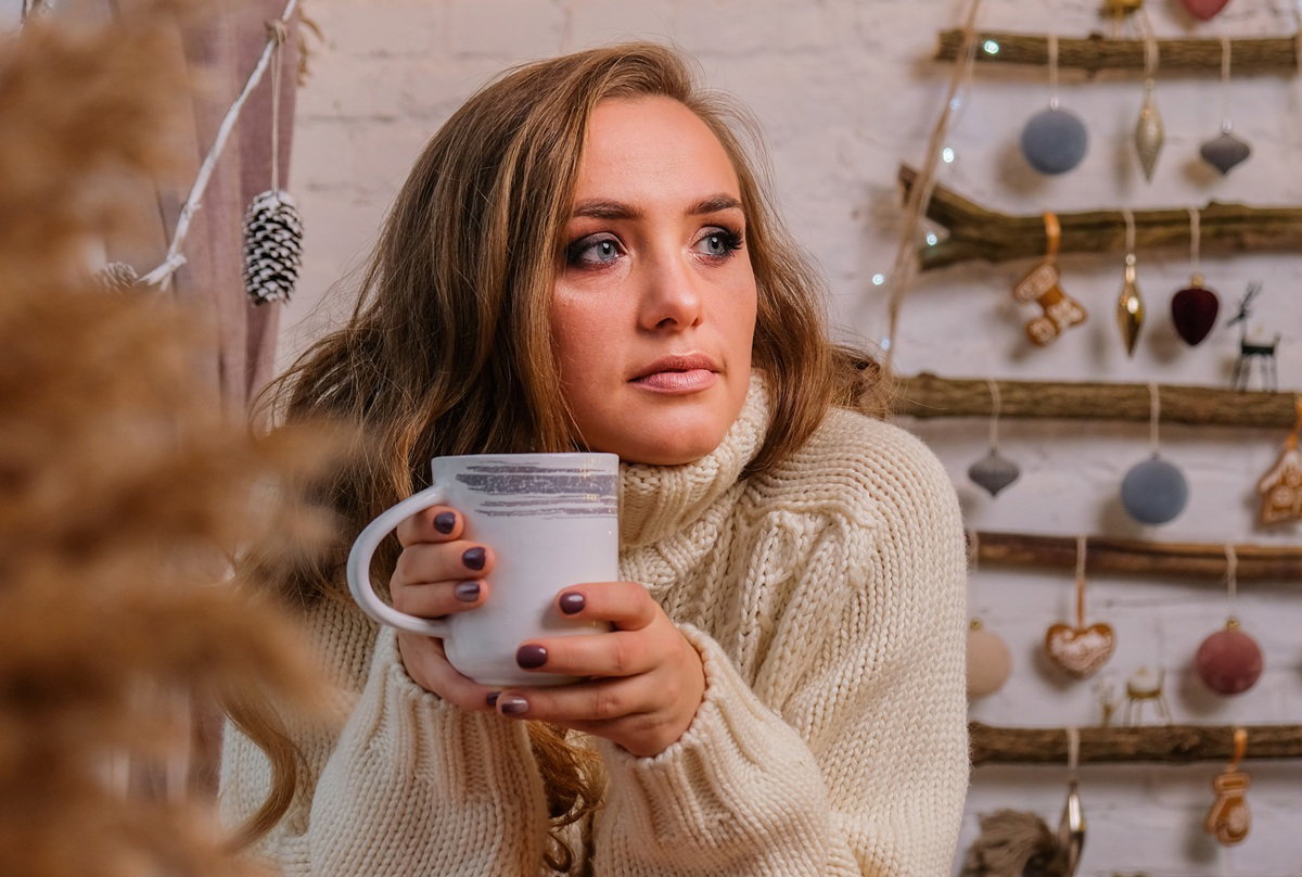 kobieta w wieku 30 plus w beżowym swetrze z kubkiem w dłoniach siedzi zamyślona