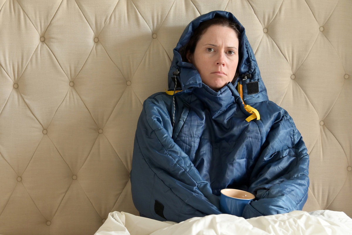 kobieta w wieku około 40 lat siedzi w łóżku okutana i z kubkiem gorącego napoju w ręku smutna