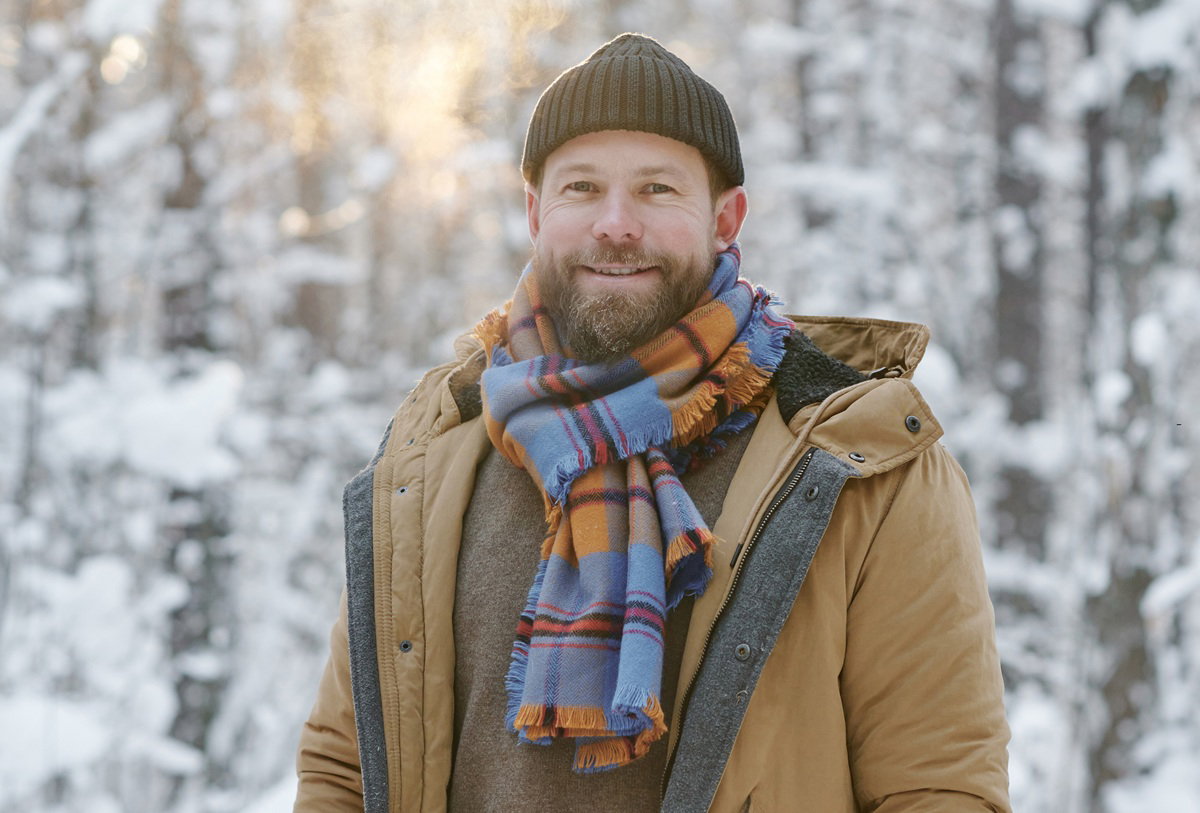 mężczyzna w wieku 35 plus brodaty stoi na tle zimowego pejzażu uśmiechnięty w czapce kurtce i kraciastym szalu