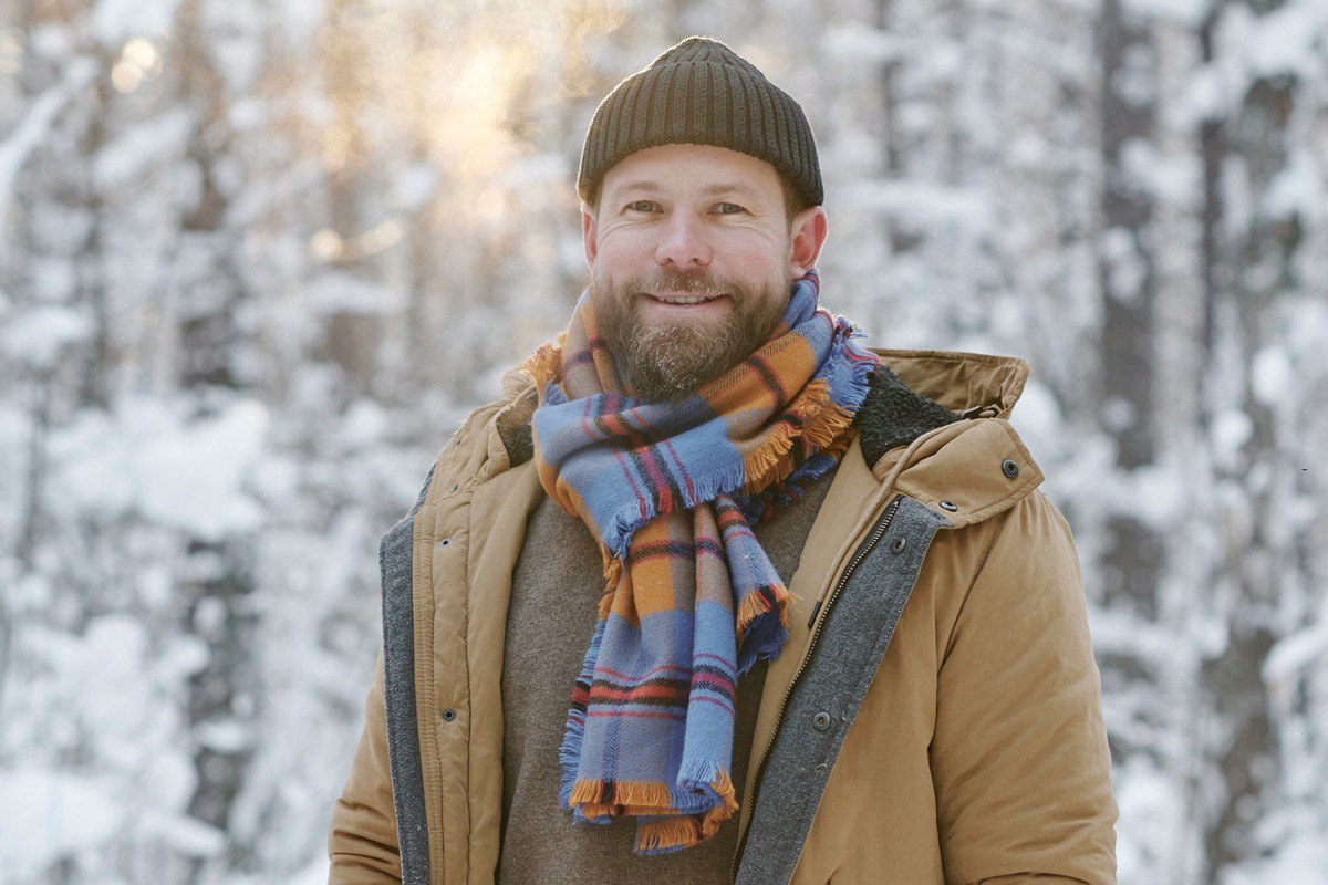 mężczyzna w wieku 35 plus brodaty stoi na tle zimowego pejzażu uśmiechnięty w czapce kurtce i kraciastym szalu