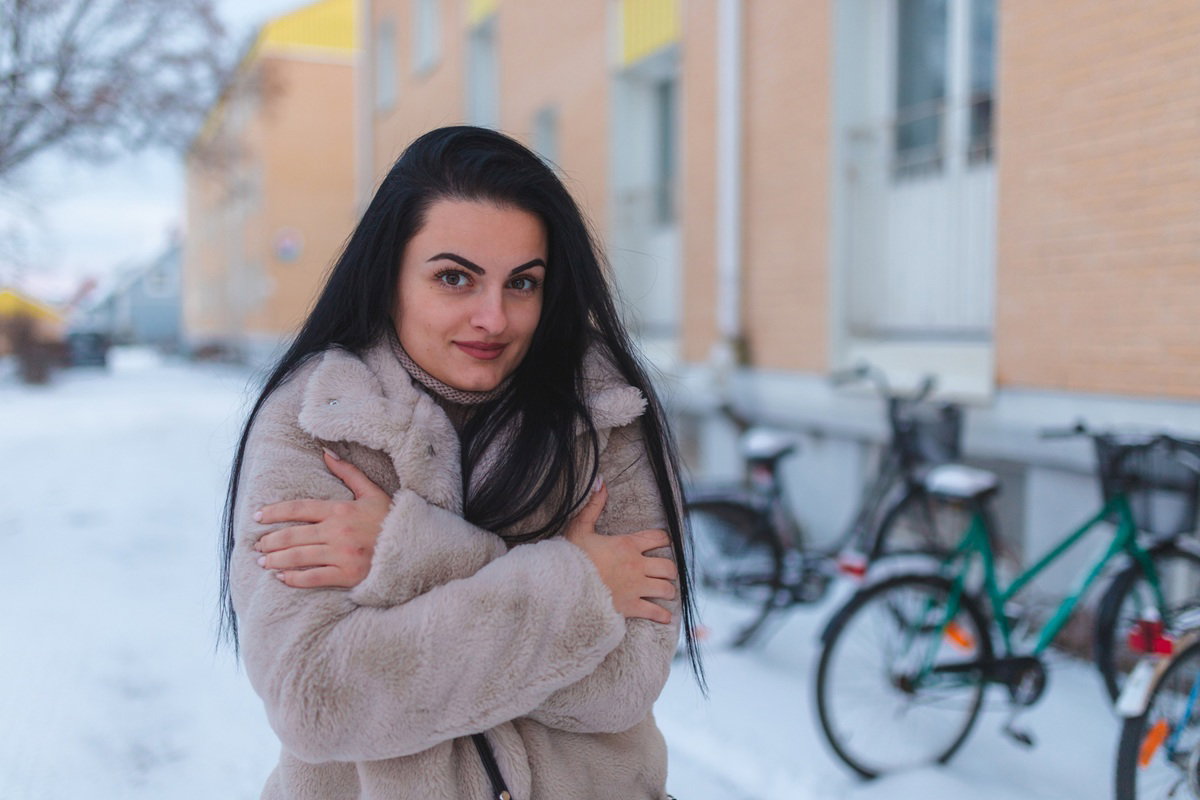 kobieta w wieku 25 plus z ciemnymi długimi włosami stoi na osiedlu i trzęsie się z zimna z lekkim uśmiechem