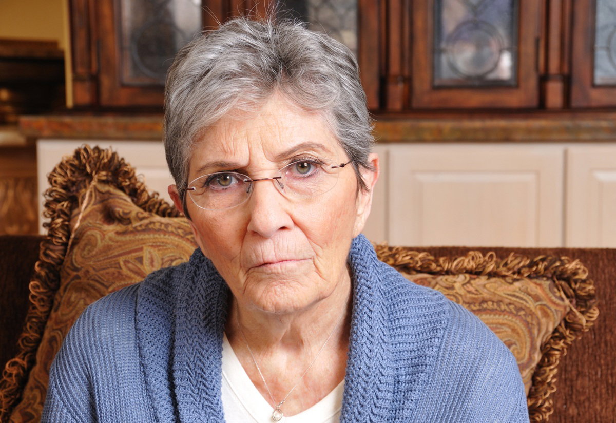 kobieta w wieku około 70 lat z krótkimi włosami i w okularach siedzi smutna i zła na kanapie