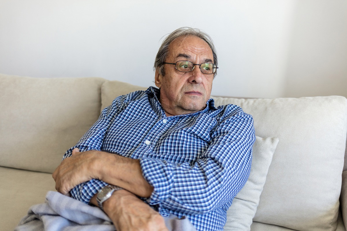 mężczyzna w wieku 70 plus w koszuli w kratę siedzi zamyślony na kanapie