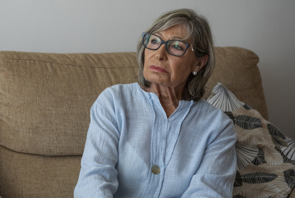 elegancka kobieta w wieku 70 plus w okularach siedzi na kanapie zamyślona i smutna