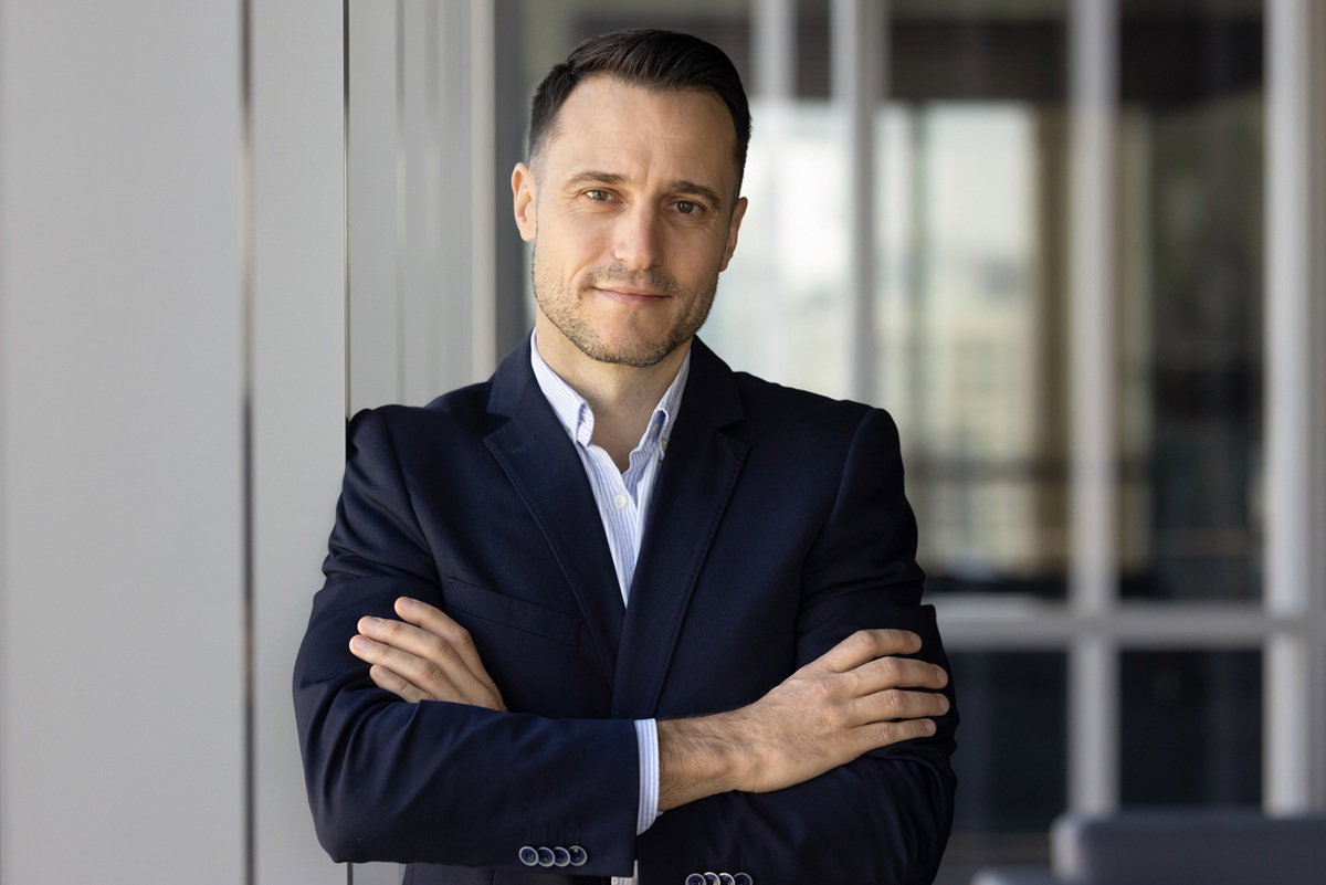 mężczyzna w wieku 35 plus stoi na tle biurowaca z założonymi rekami i ma smutny uśmiech