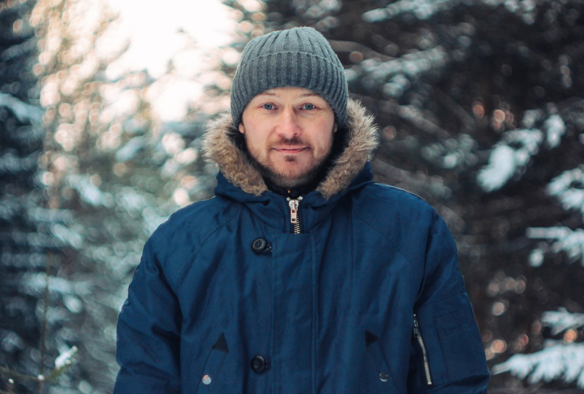 mężczyzna w wieku 35 plus stoi na zewnątrz na tle zaśnieżonych drzew i lekko się uśmiecha