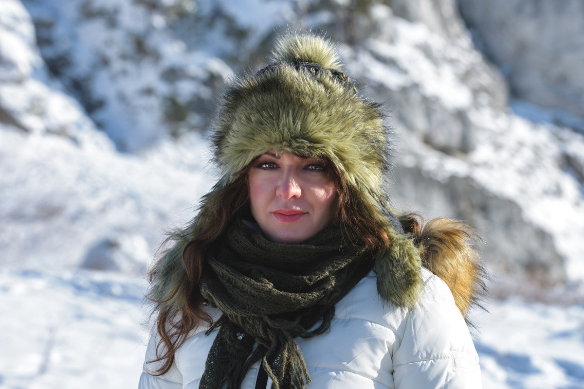 kobieta w wieku 35 plus stoi w futrzanej czapie na tle gór z poważnym wyrazem twarzy