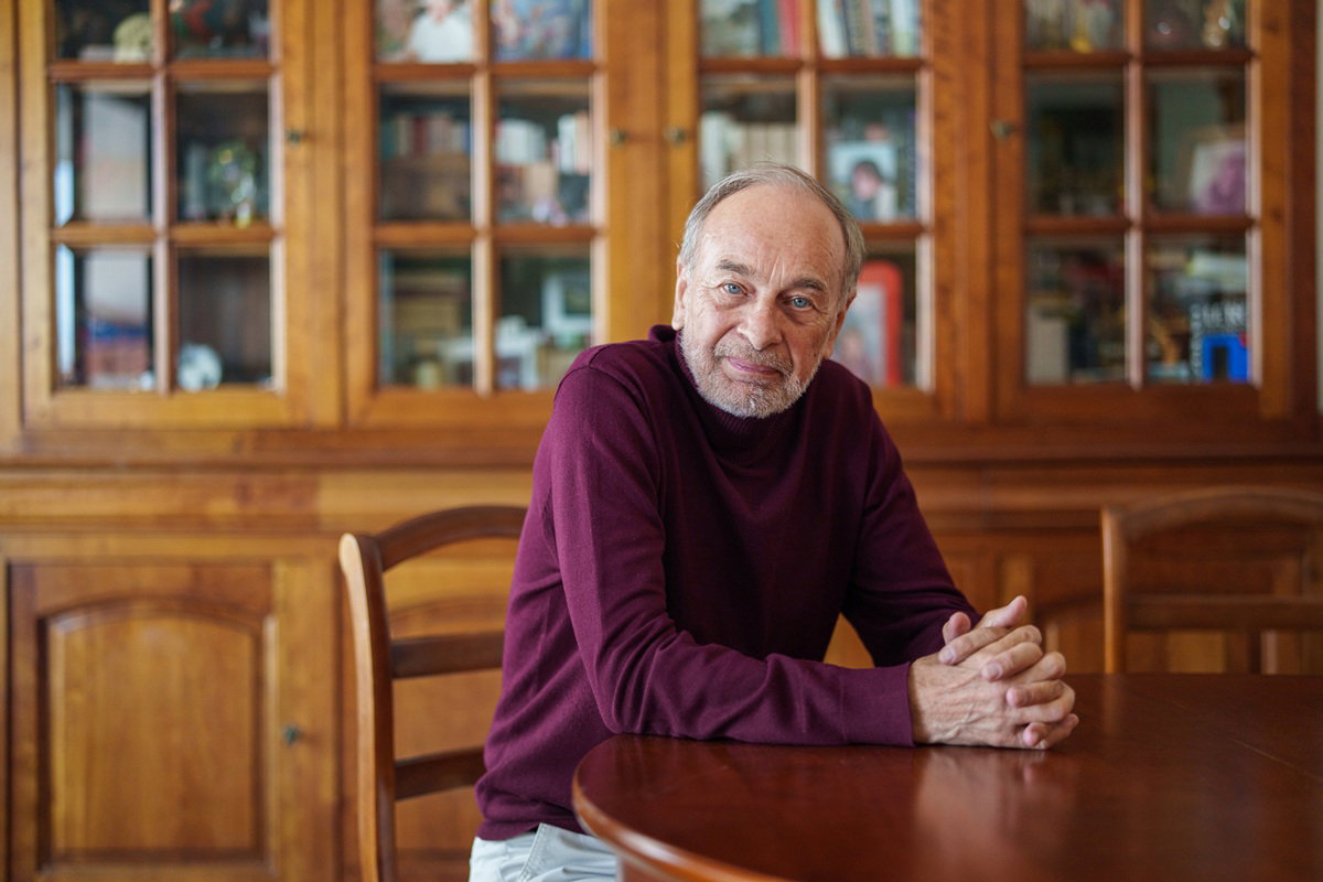 mężczyzna w wieku 70 plus w bordowym golfie siedzi przy stole smutny na tle mebli