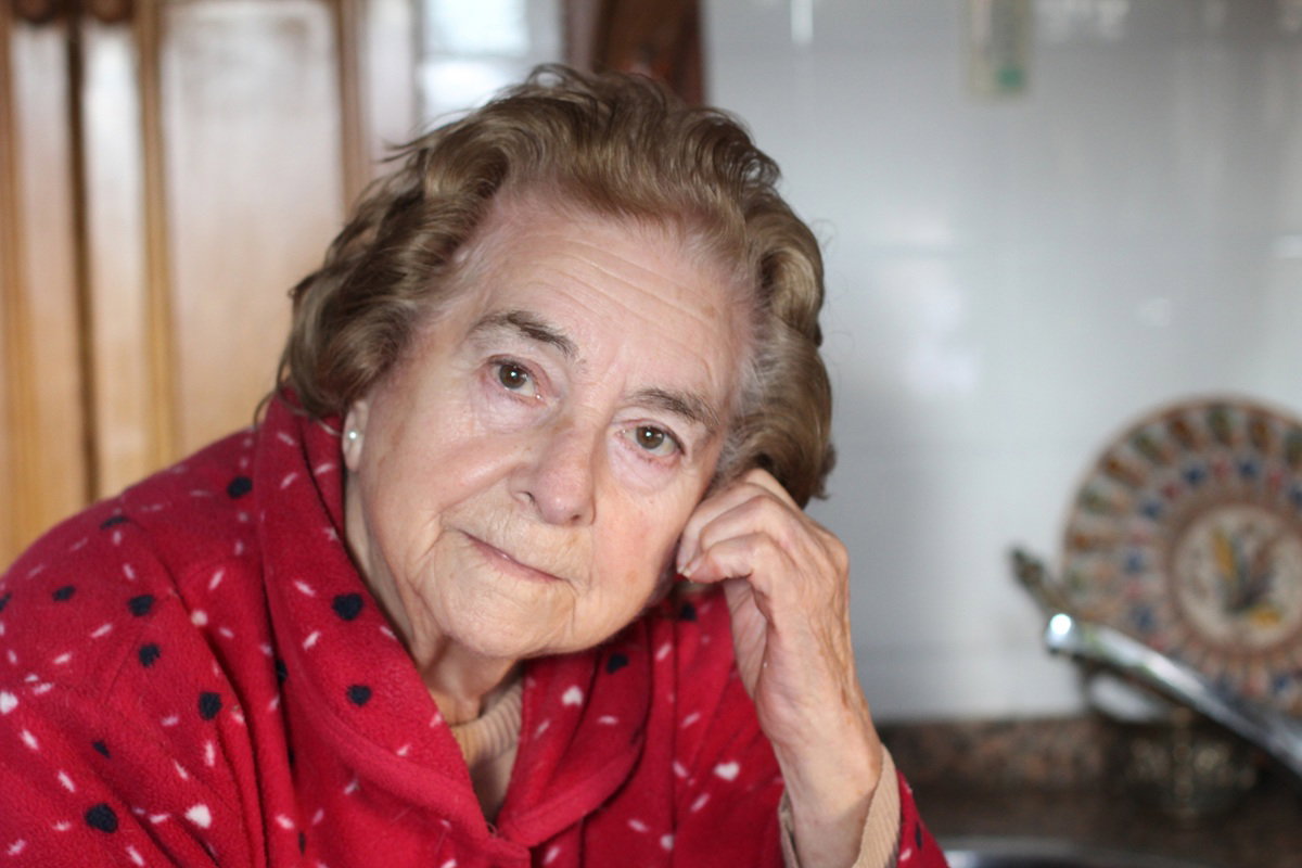 kobieta w wieku około 70 lat w czerwonym szlafroku siedzi zamyślona i smutna w kuchni