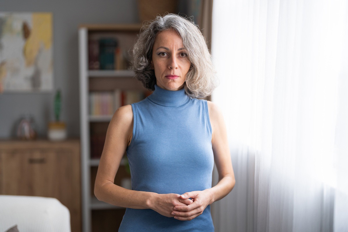 kobieta w wieku 40 plus z siwymi włosami w niebieskiej sukience stoi zamyślona i smutna