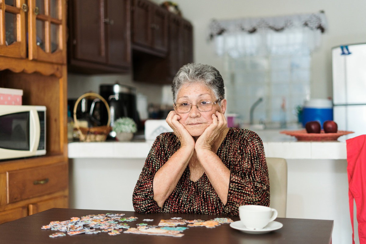 kobieta w wieku 70 plus w okularach siedzi w kuchni zasępiona przy stole z filiżanką i nad puzzlami