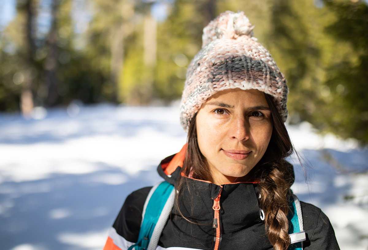kobieta w wieku 30 plus w czapce z pomponem stoi na tle zimowego pejzażu z lekkim uśmiechem