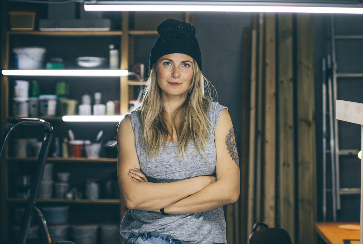 kobieta w wieku 35 plus w czarnej czapce i szarej koszulce stoi w piwnicy ze skrzyżowanymi ramionami zamyślona