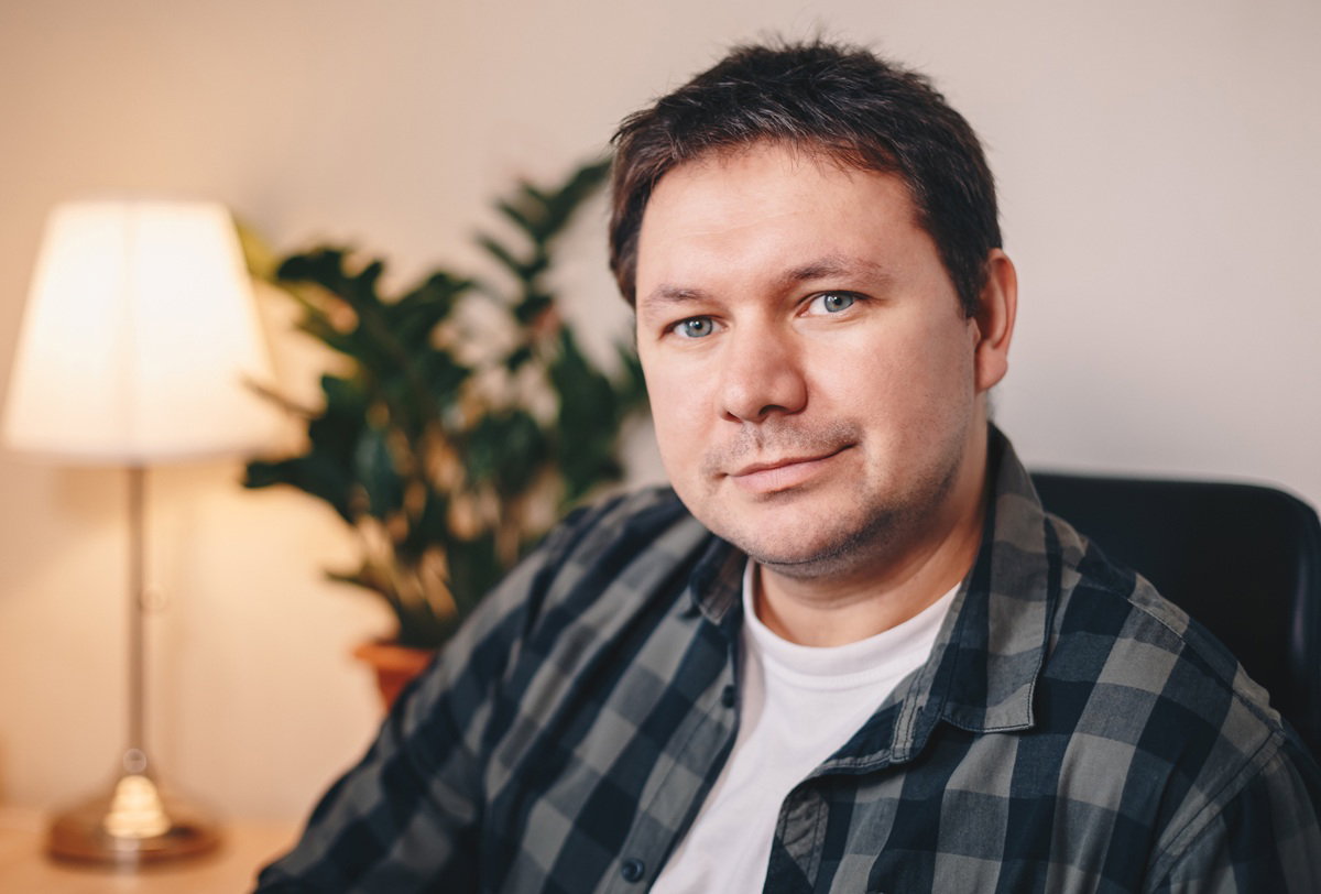 mężczyzna w wieku 35 plus w kracistej koszuli siedzi w fotelu i lekko się uśmiecha