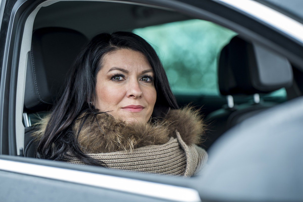 kobieta w wieku 35 plus z ciemnymi długimi włosami siedzi w samochodzie w kurtce i patrzy przenikliwym wzrokiem
