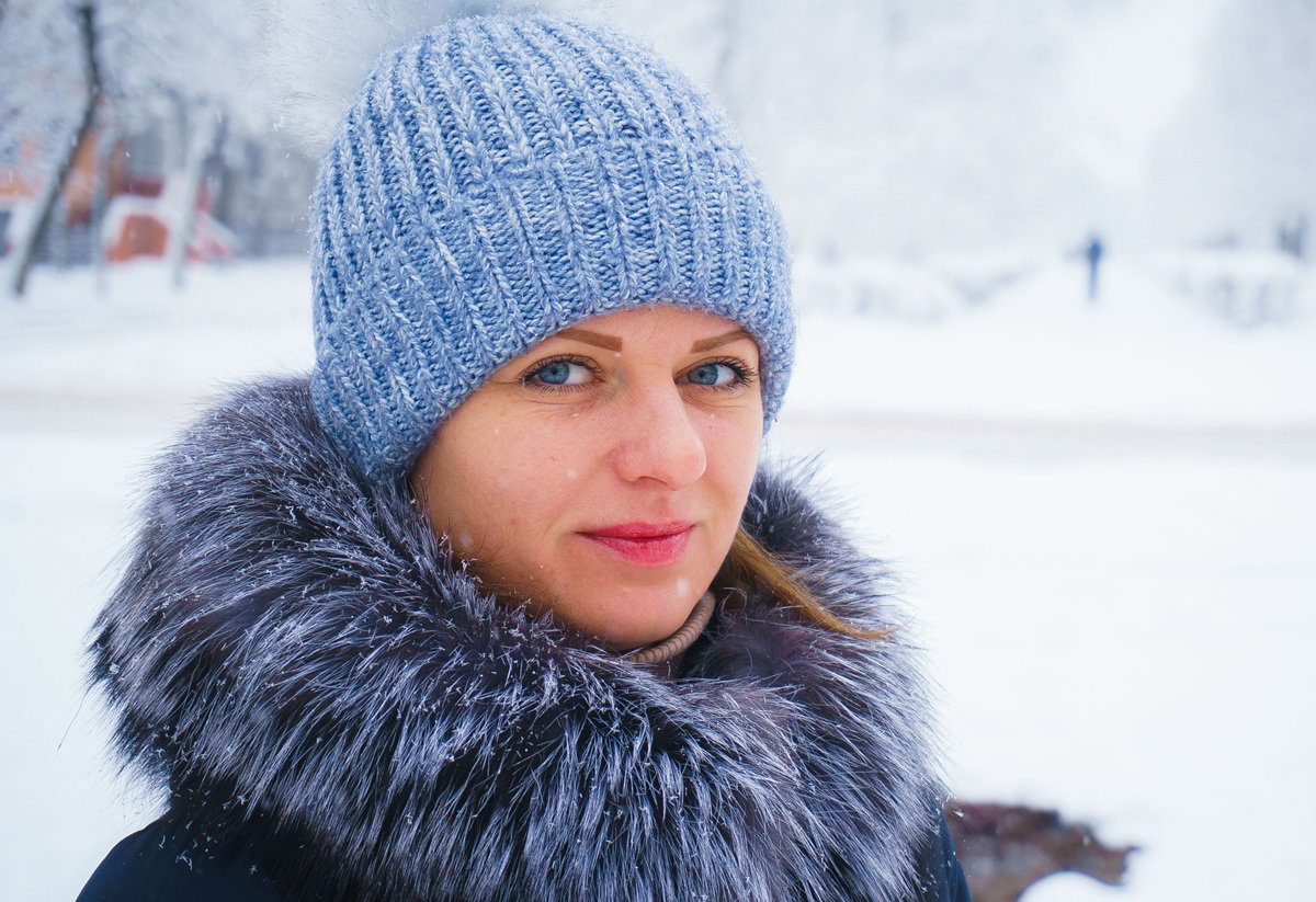 kobieta w wieku 25 plus w czapce i w kurtce z futrzanym kołnierzem stoi na zewnątrz na tle zimowego pejzażu