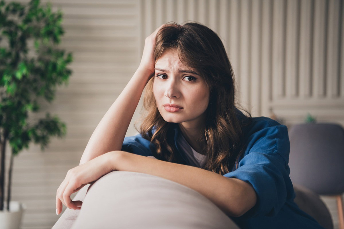 kobieta w wieku 25 plus z długimi włosami i niebieskiej bluzce siedzi na kanapie smutna i zamyślona