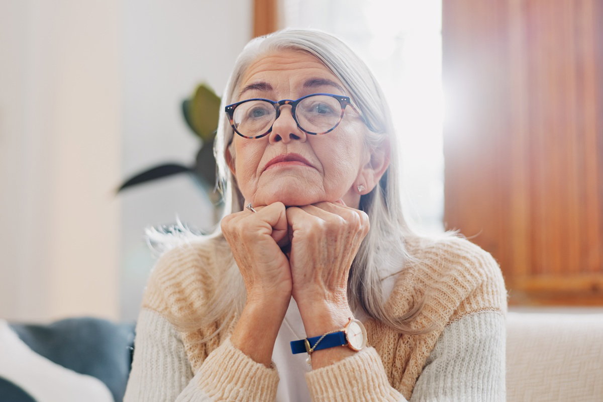 kobieta w wieku 65 plus w okularach z siwymi włosami siedzi na tle jasnego okna zamyślona