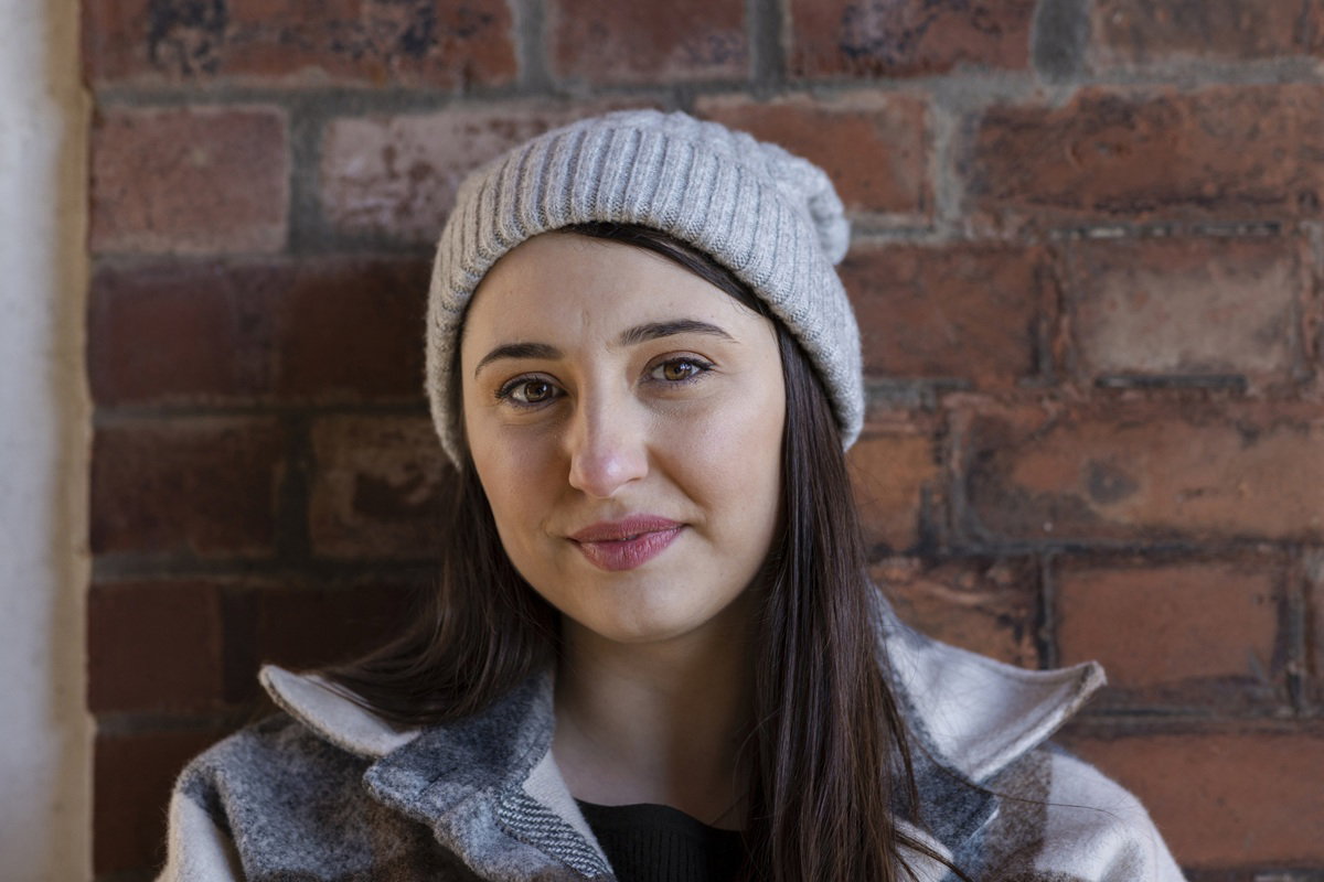 kobieta w wieku 30 plus w czapce stoi na tle ściany z cegieł z leciutkim uśmiechem wzruszona