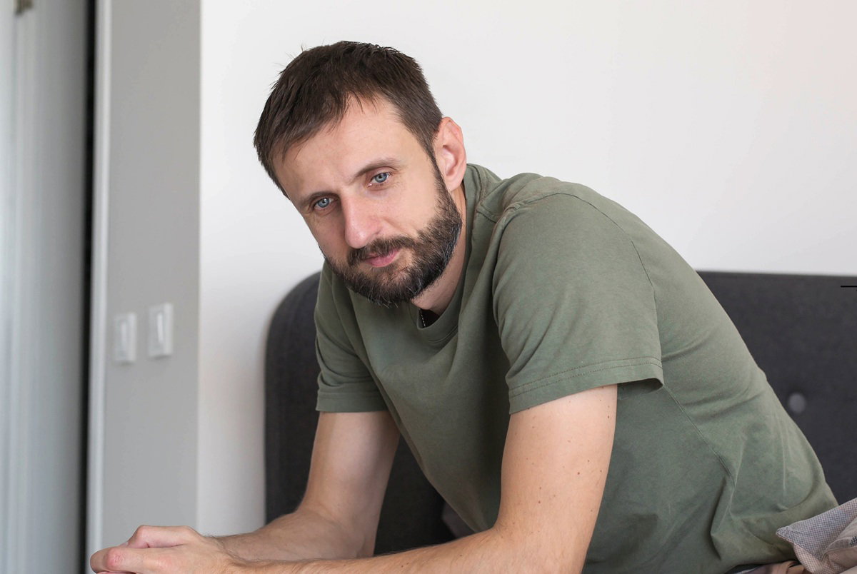 mężczyzna w wieku 35 plus z wąsami i brodą w zielonej koszulce siedzi na łóżku zamyślony i zmartwiony