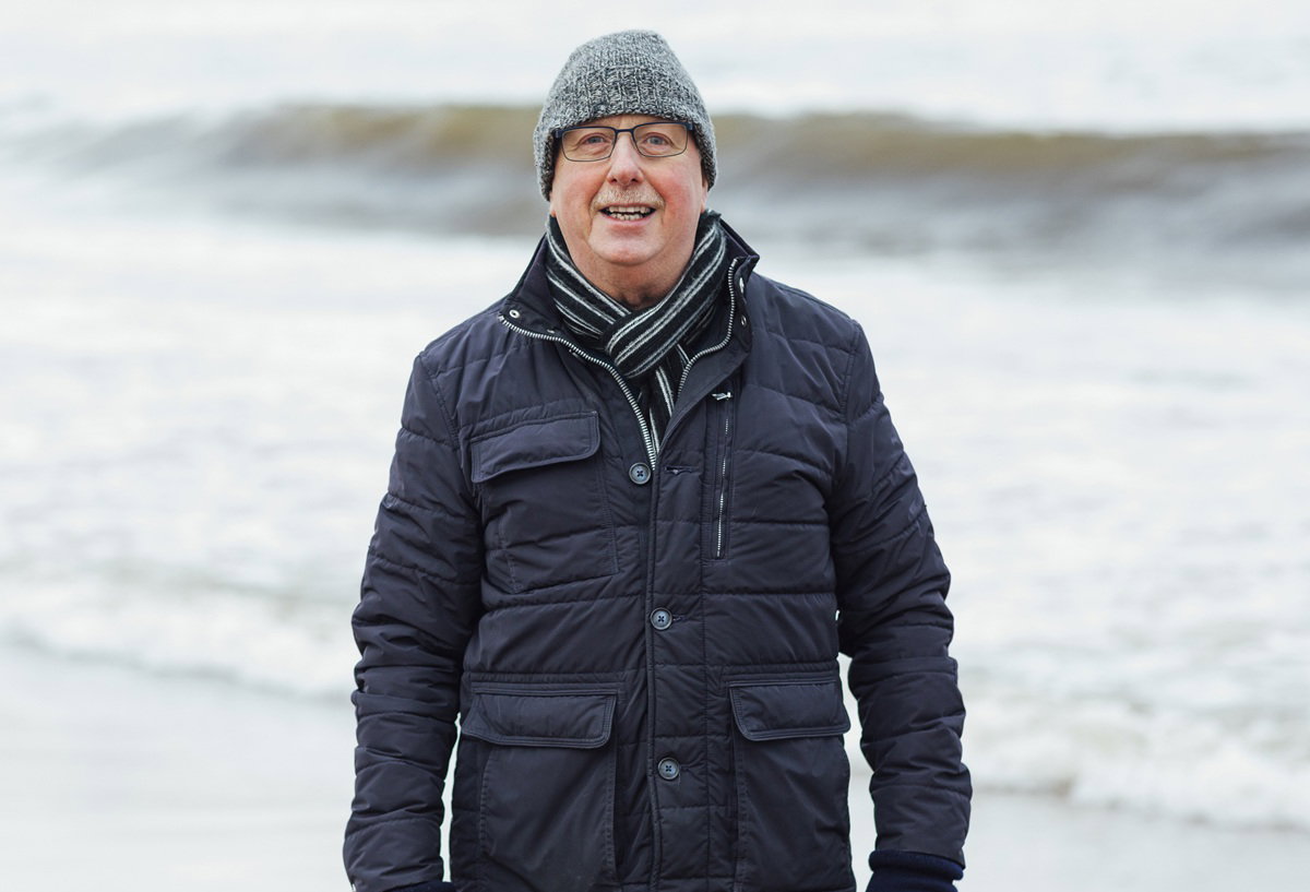 mężczyzna w wieku 65 plus w granatowej kurtce stoi na tle morza i się uśmiecha